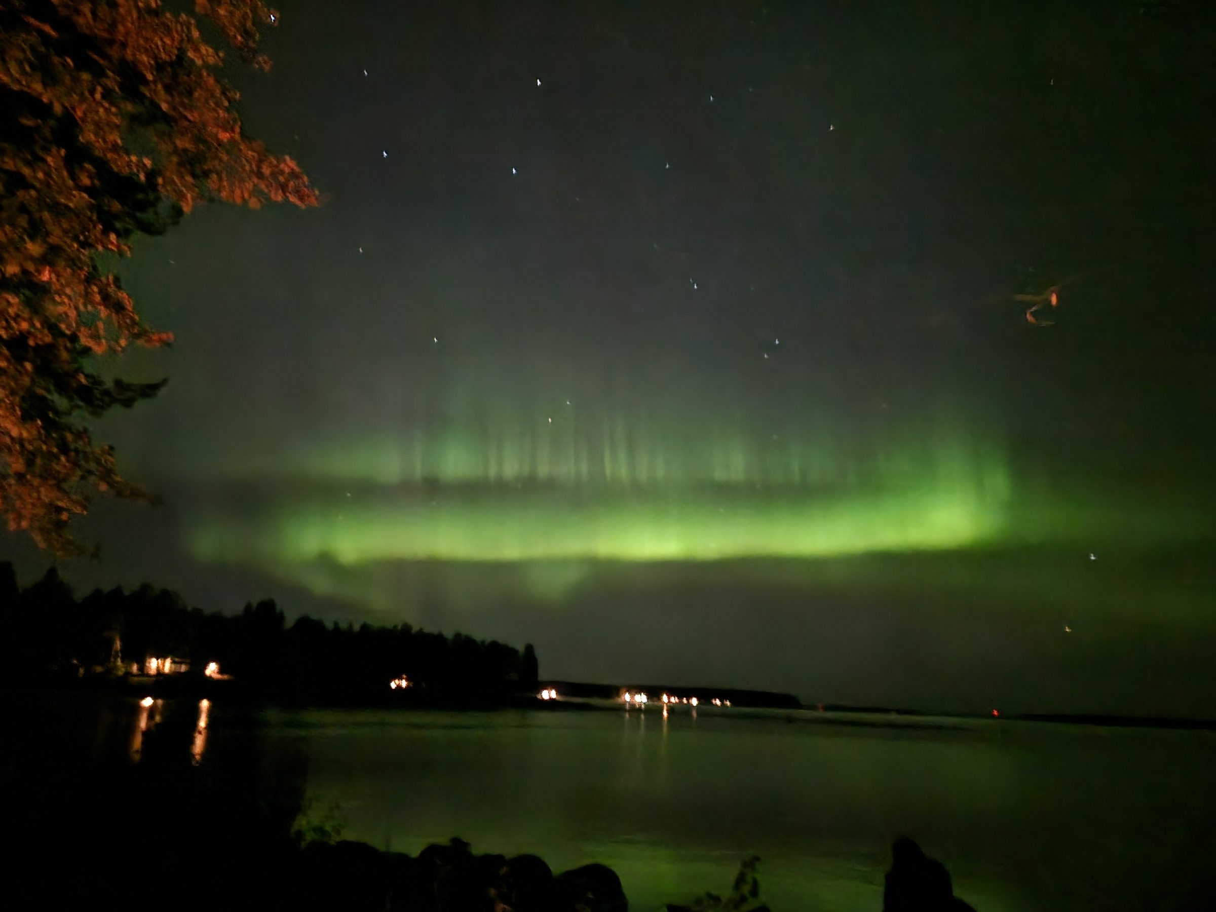 A bright green aurora band stretched low across the sky and mirrored in a still Lapland lake, with shoreline lights below.