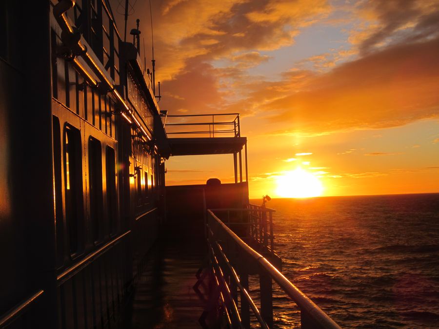Golden sunset over the sea viewed along the side railings and deck of an expedition ship.