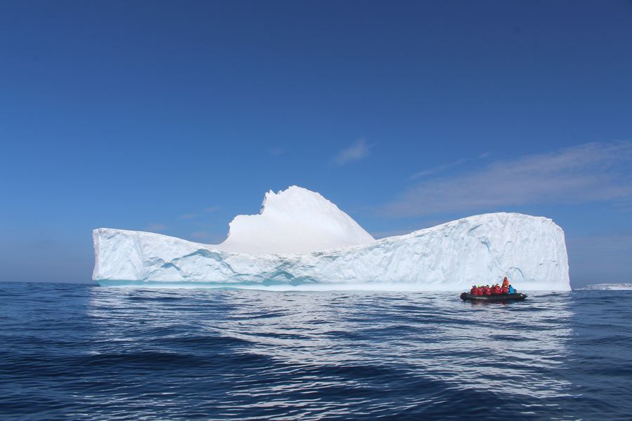 Small inflatable zodiac with passengers beside a large white iceberg on a calm dark ocean under blue sky.
