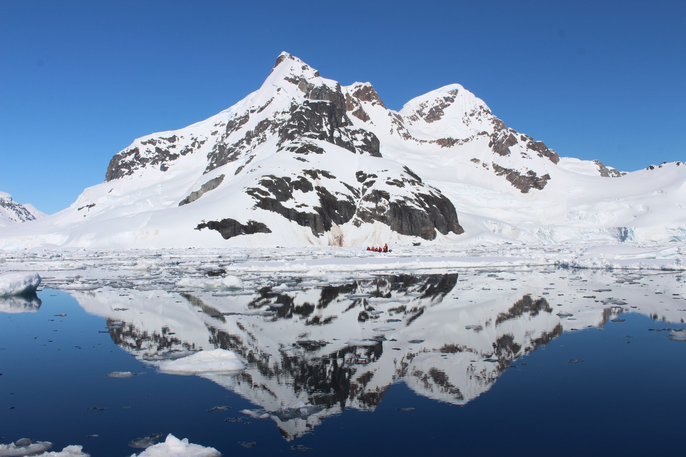 Snow-covered jagged mountain mirrored in still dark water under a deep blue sky, with floating ice.