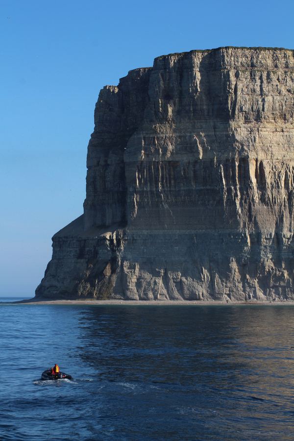 Towering vertical sea cliff rising from calm water with a tiny boat below for scale.