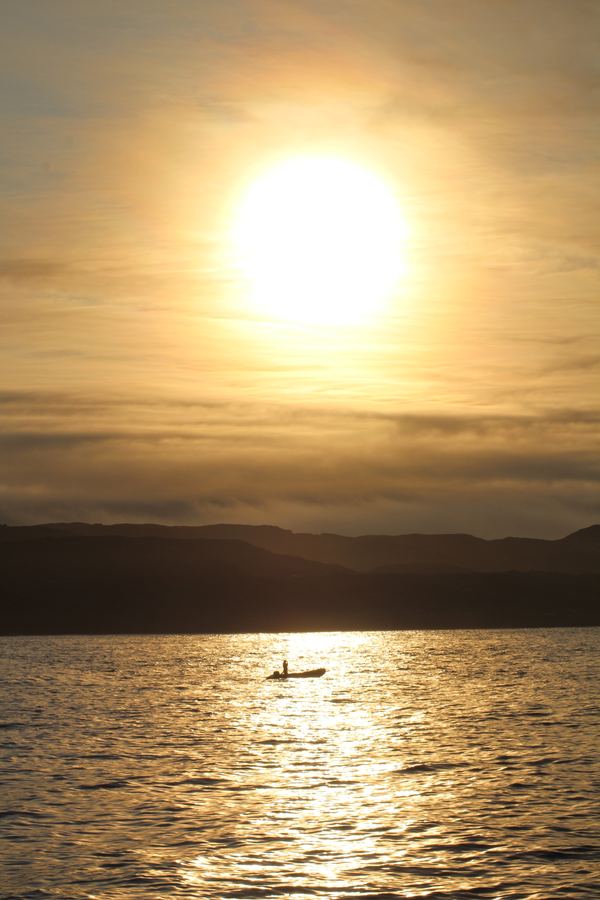 Low golden sun over calm Arctic water with a lone kayaker in silhouette.