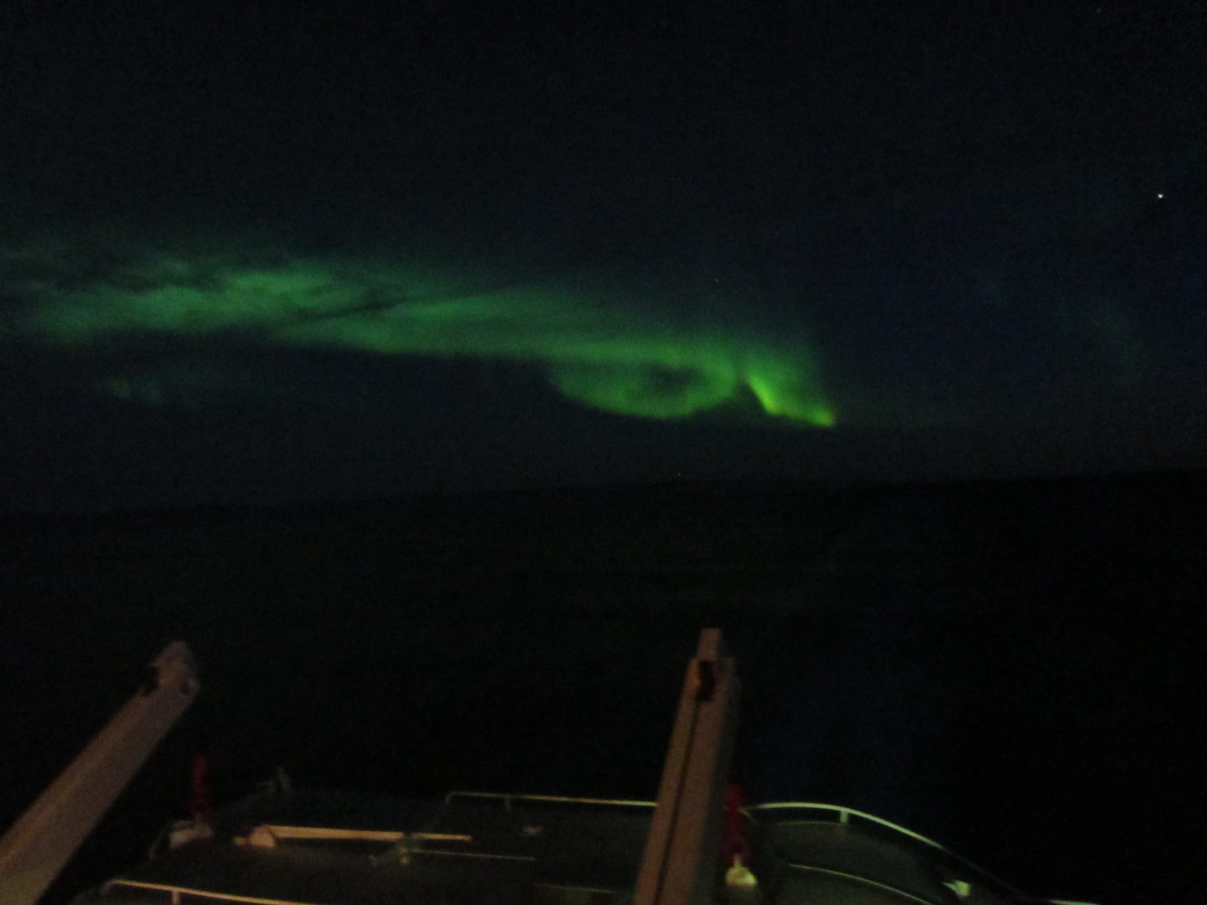 Bright green aurora arcing across a dark night sky over a ship railing in the foreground.