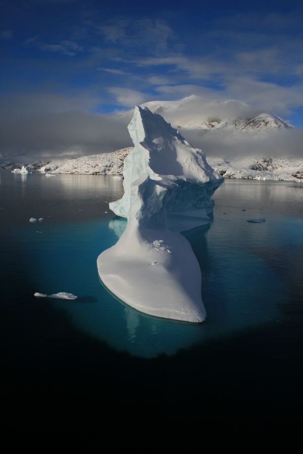 Tall sculptural white iceberg with a turquoise underwater base under a blue sky.