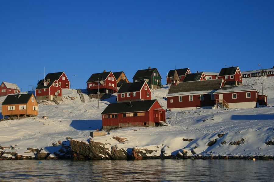 Red and yellow wooden houses on a snowy rocky shore beside calm water.
