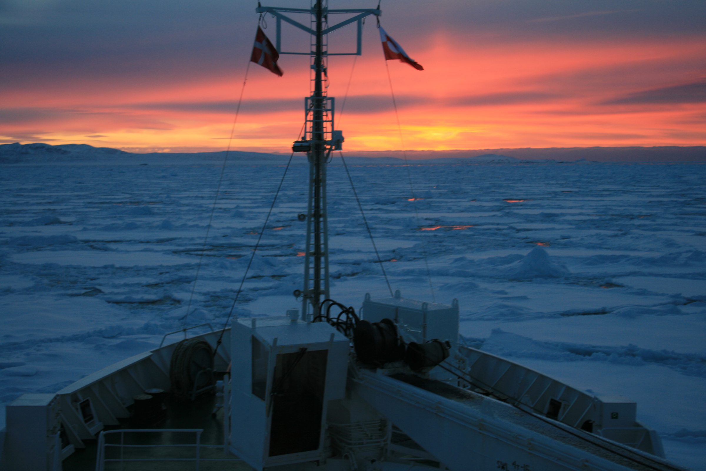 Ship's bow pushing through pack ice under a glowing orange and red twilight sky over the sea.