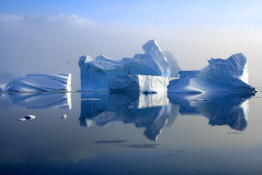 Large pale-blue iceberg with sculpted ridges and a clear mirror reflection on still water.