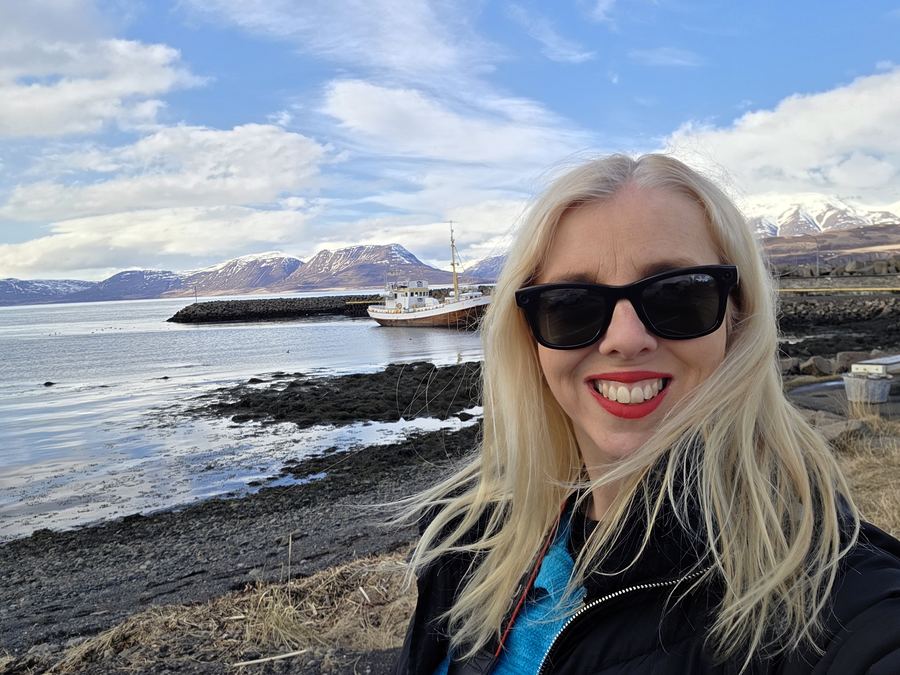 Smiling blonde woman in sunglasses at a coastal harbour with a boat and snowy hills behind.