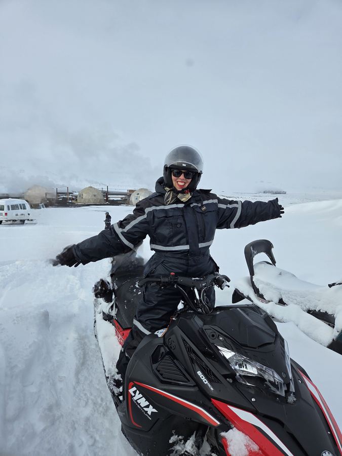 Person in black snowsuit and helmet sitting on a red and black snowmobile in deep snow, arms out.