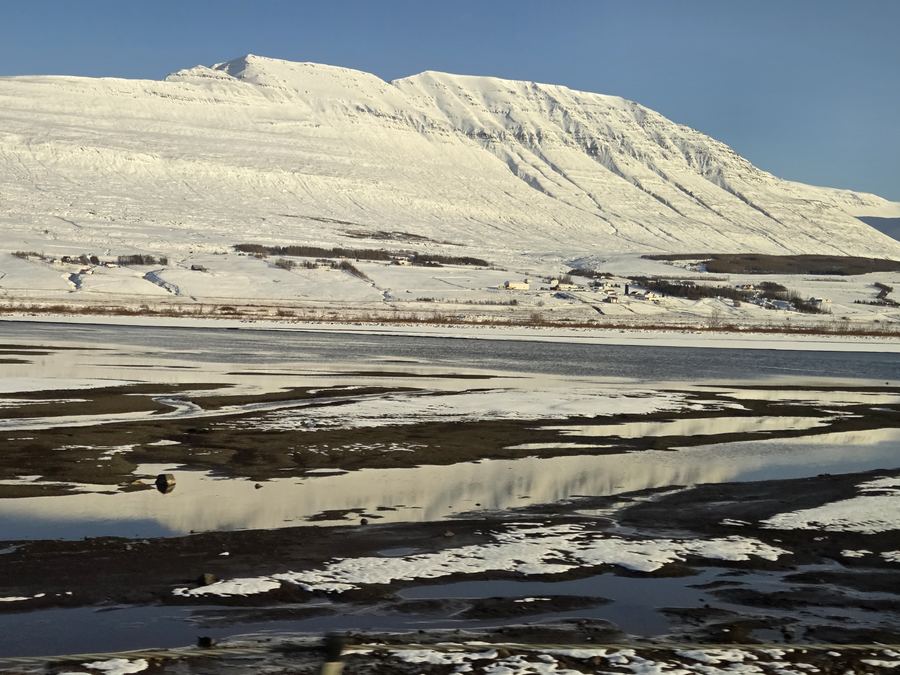 Snow-covered mountain ridge reflected in still water amid patches of snow and dark ground.