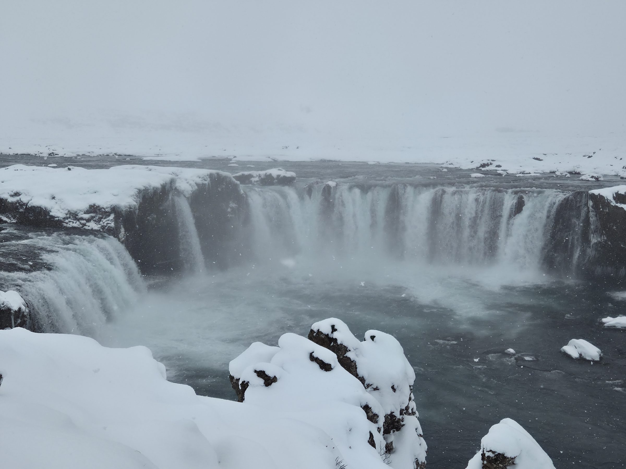Wide waterfall pouring over a rock ledge into a river, snow-covered banks and mist under grey sky.