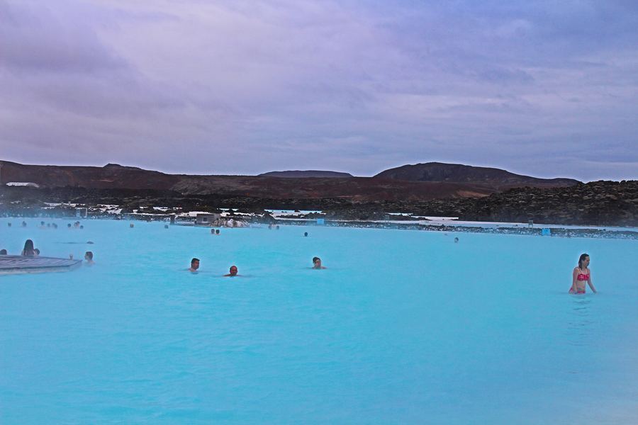 People bathing in a bright turquoise geothermal lagoon below low hills and a pale sky.