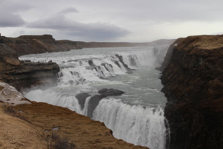 Wide tiered waterfall cascading over rock ledges into a canyon under a heavy grey sky.