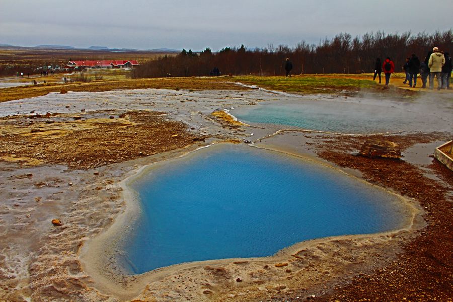 Vivid blue hot spring pool ringed by rust-orange mineral crust with visitors in the distance.