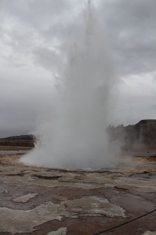 Tall column of water and steam erupting from a geyser over a mineral flat under grey clouds.