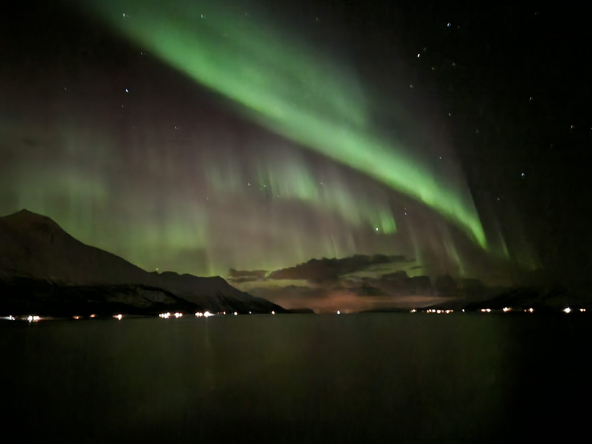 A sweeping arc of green aurora brightening over a mountain ridge above a fjord.