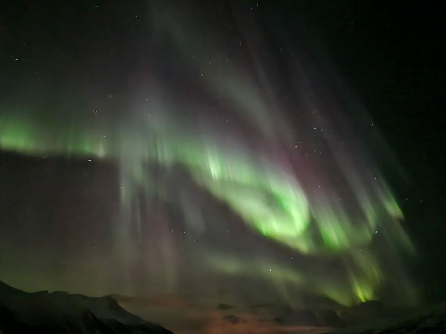 Vivid green aurora rays streaming down across the night sky above a dark mountain horizon.