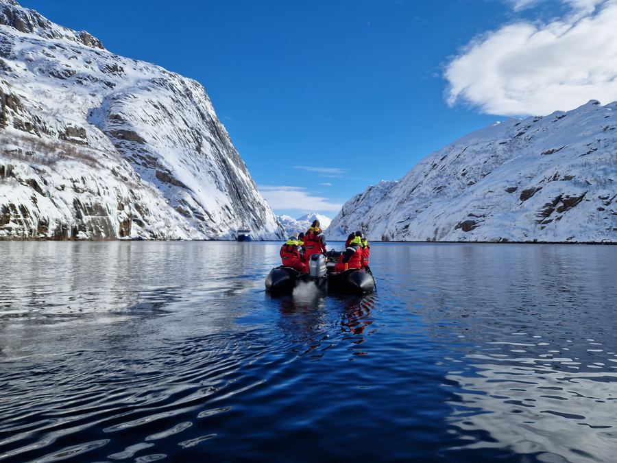 Small boat with people crossing calm blue water between steep snow-covered Arctic mountains.