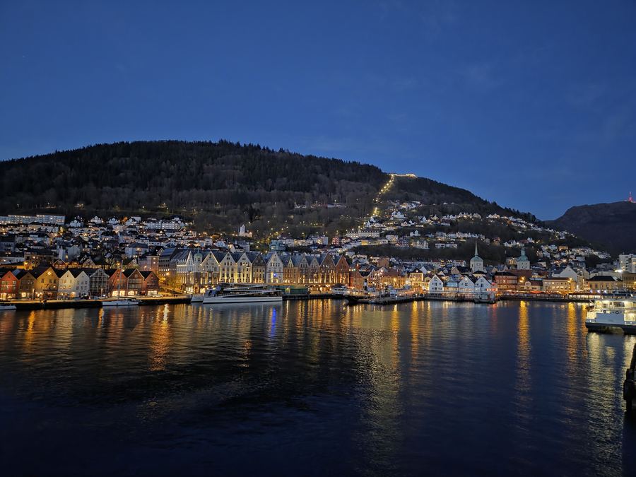 Harbour-town lights glowing at dusk beneath a dark mountain ridge and deep blue sky.