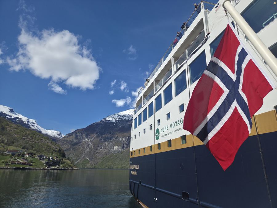 A Norwegian flag flying from a ship's stern above a calm fjord and green mountains.