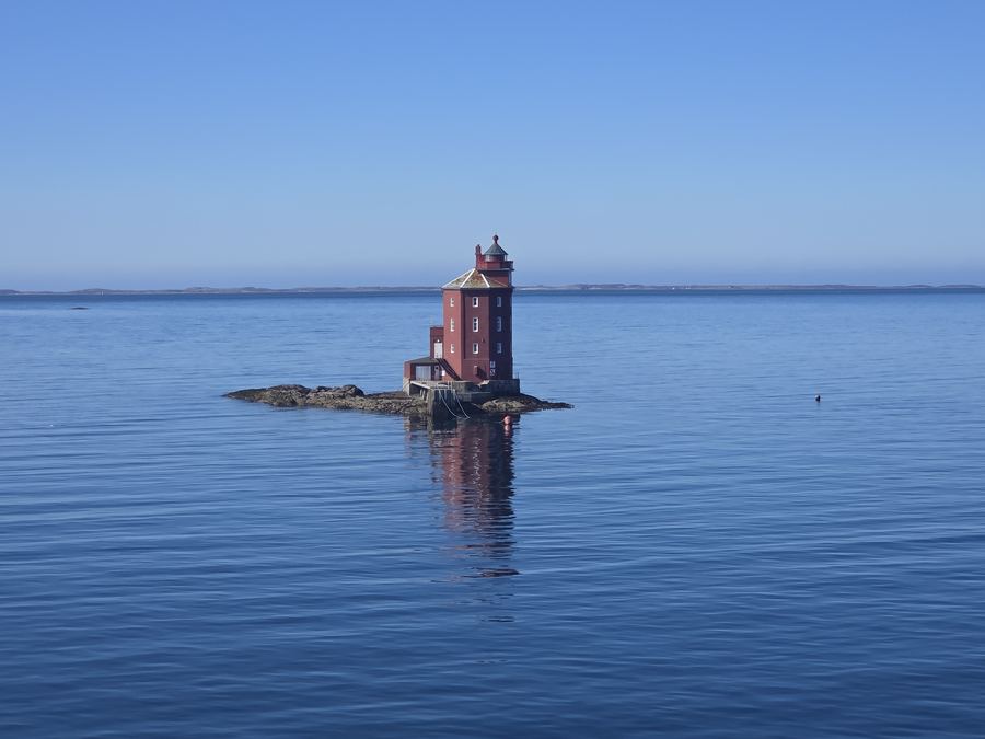 A small red lighthouse on a rocky islet in a calm sea with distant snowy mountains.