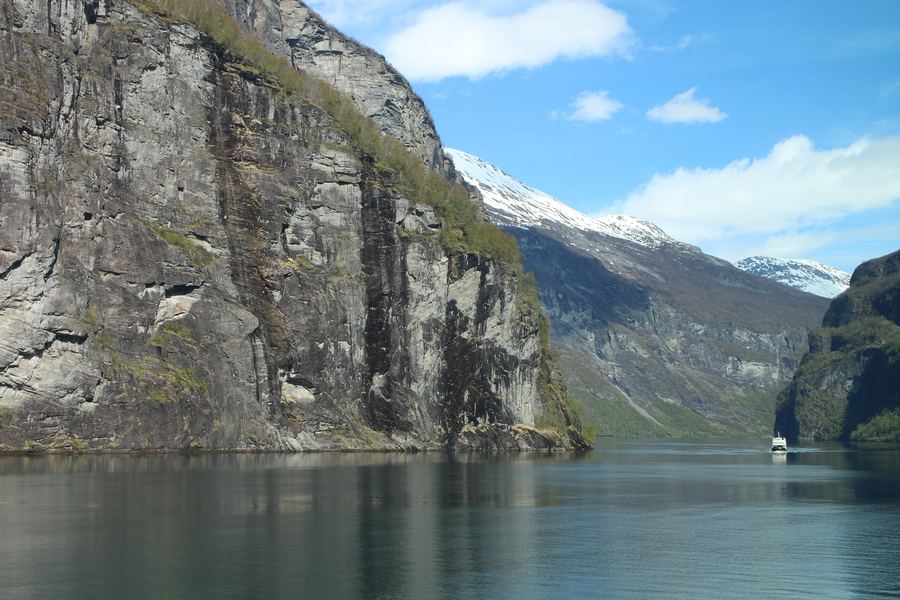 Sheer rock cliffs dropping into a turquoise fjord with a small ship passing below.
