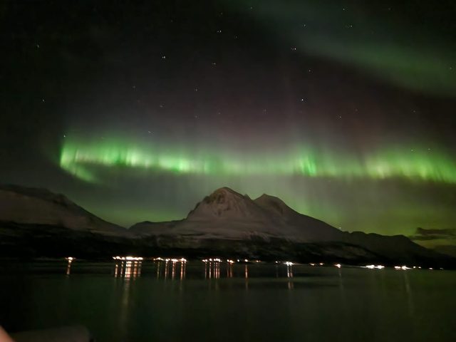 A bright green aurora band stretched across the sky above a pointed peak, town lights reflected in the fjord below.