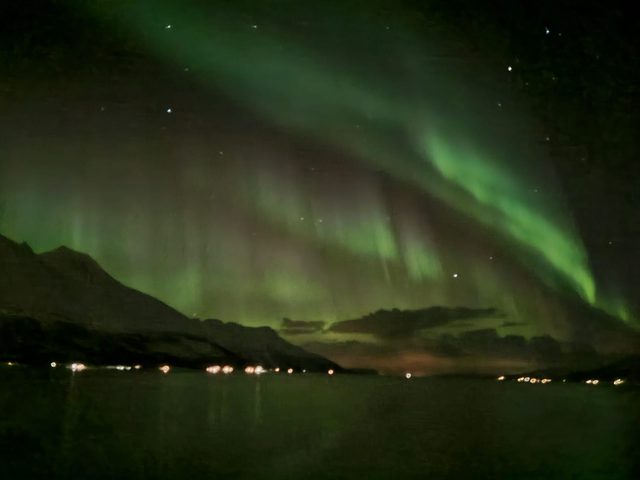 Green aurora over a fjord ringed by mountains, with town lights along the far shore.