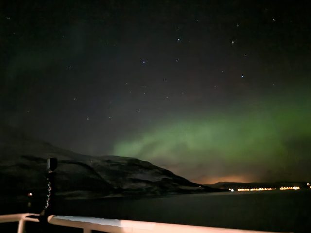Green aurora over the mountains, seen from the deck rail of a coastal ship.