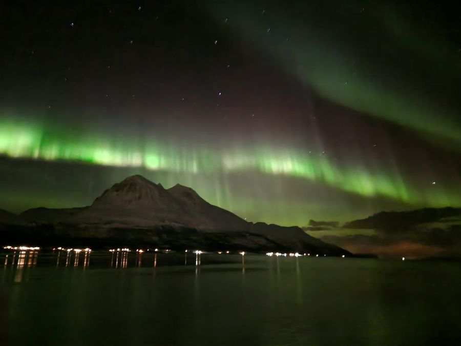 Bright green aurora curtains over a fjord ringed by snow-dusted mountains and shore lights.