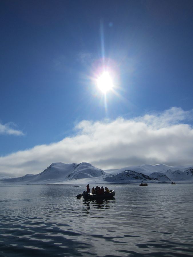 Bright sun over calm water with a small boat and distant snow-covered mountains.