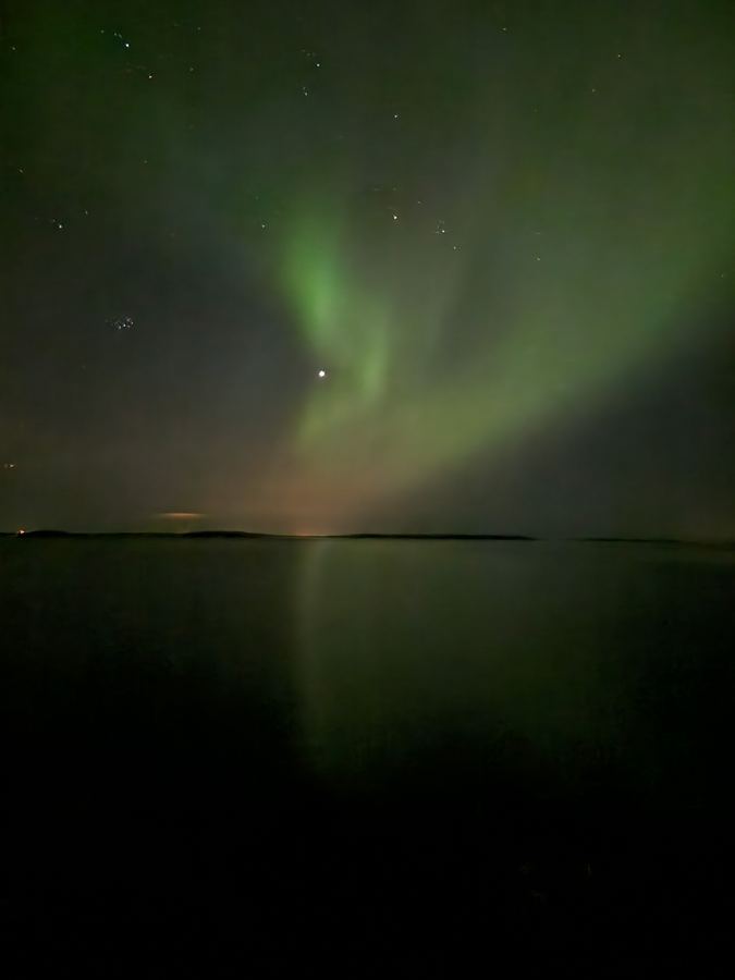 Soft green aurora glow above a lake with a faint reflection on the dark water.