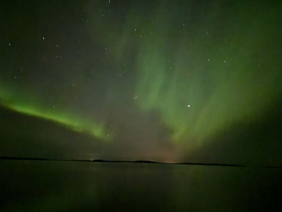 Broad green aurora sweeping across a starlit sky over water with a low dark horizon.