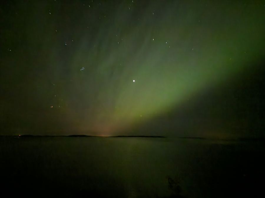 Wide aurora with faint pink and green tones over a calm horizon and dark water.